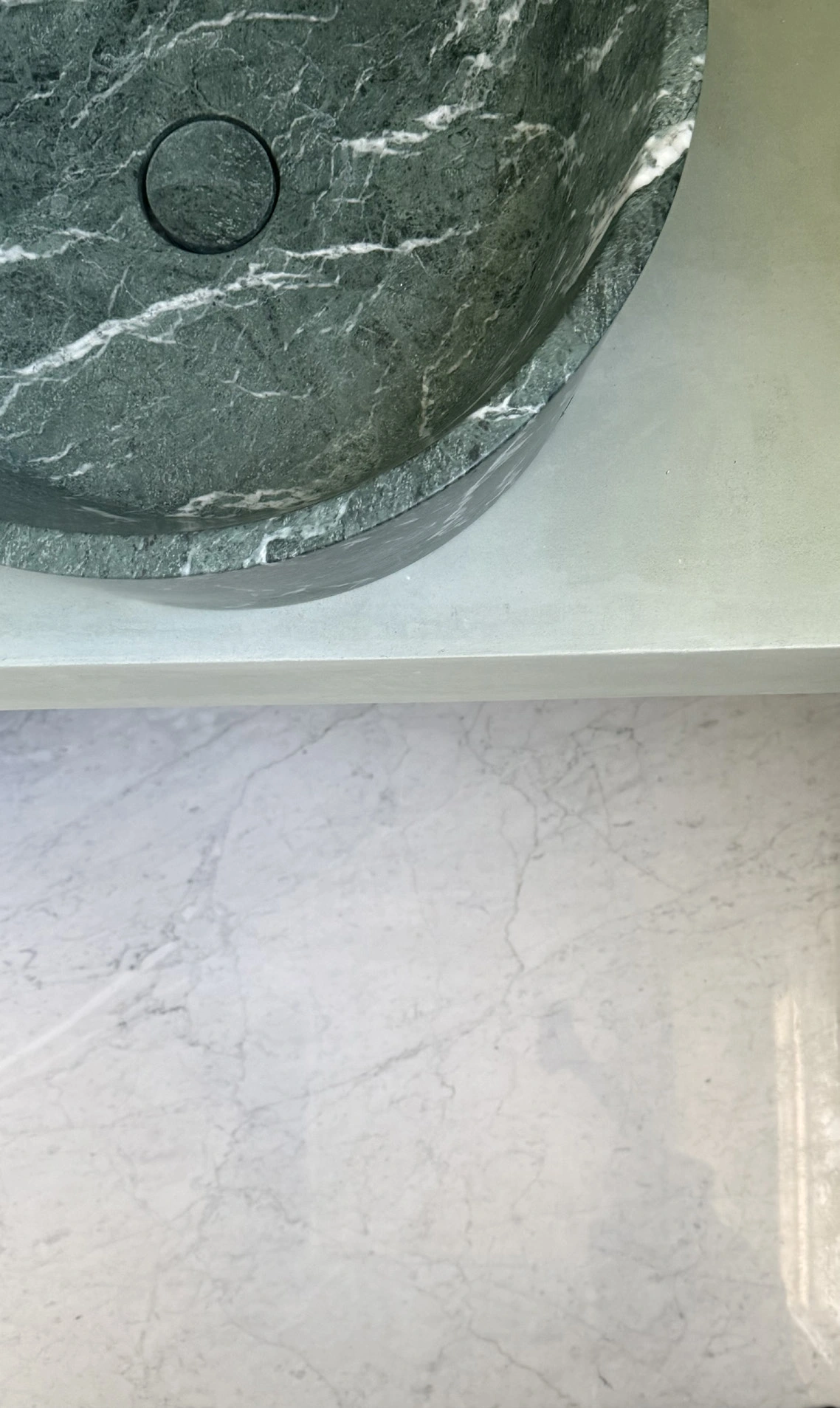 Close detail of a dark green marble vessel basin above a pale stone vanity, with a circular waste fitting.