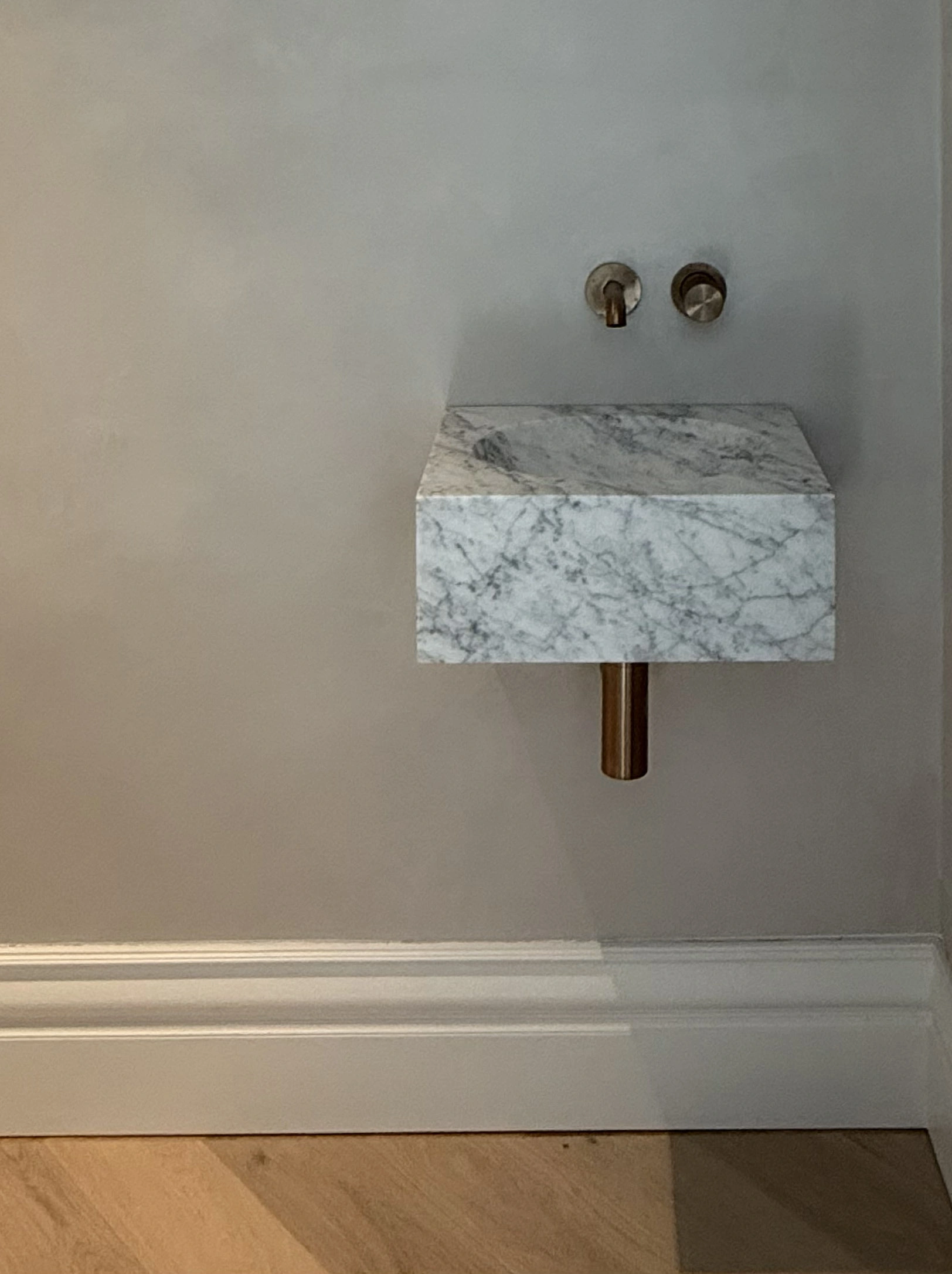 Compact floating white-and-grey marble wall basin with brass wall-mounted taps against a pale plaster wall.