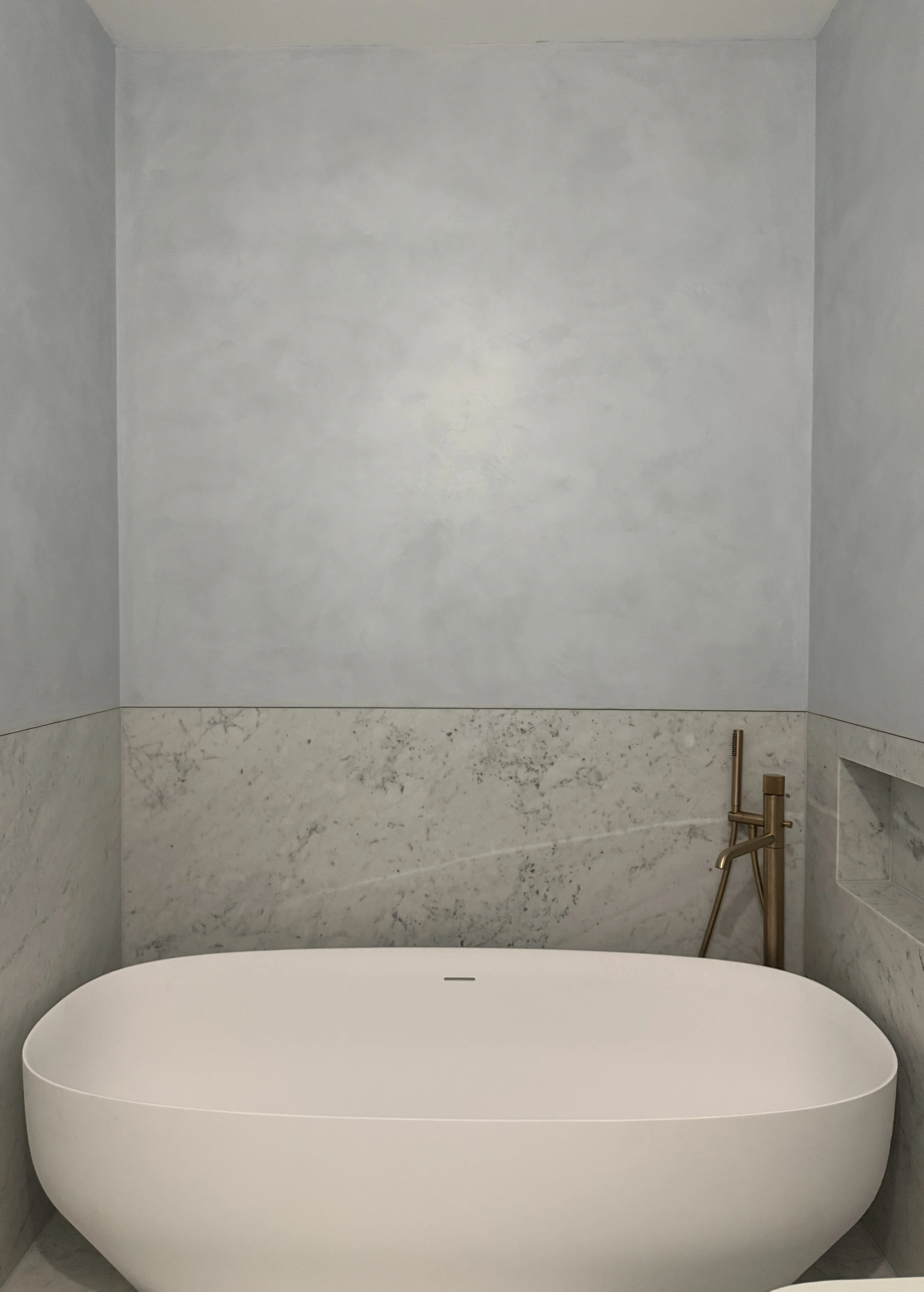 Freestanding white oval bath set against pale Venetian plaster and a grey-veined marble surround, with brass floor-mounted fittings.