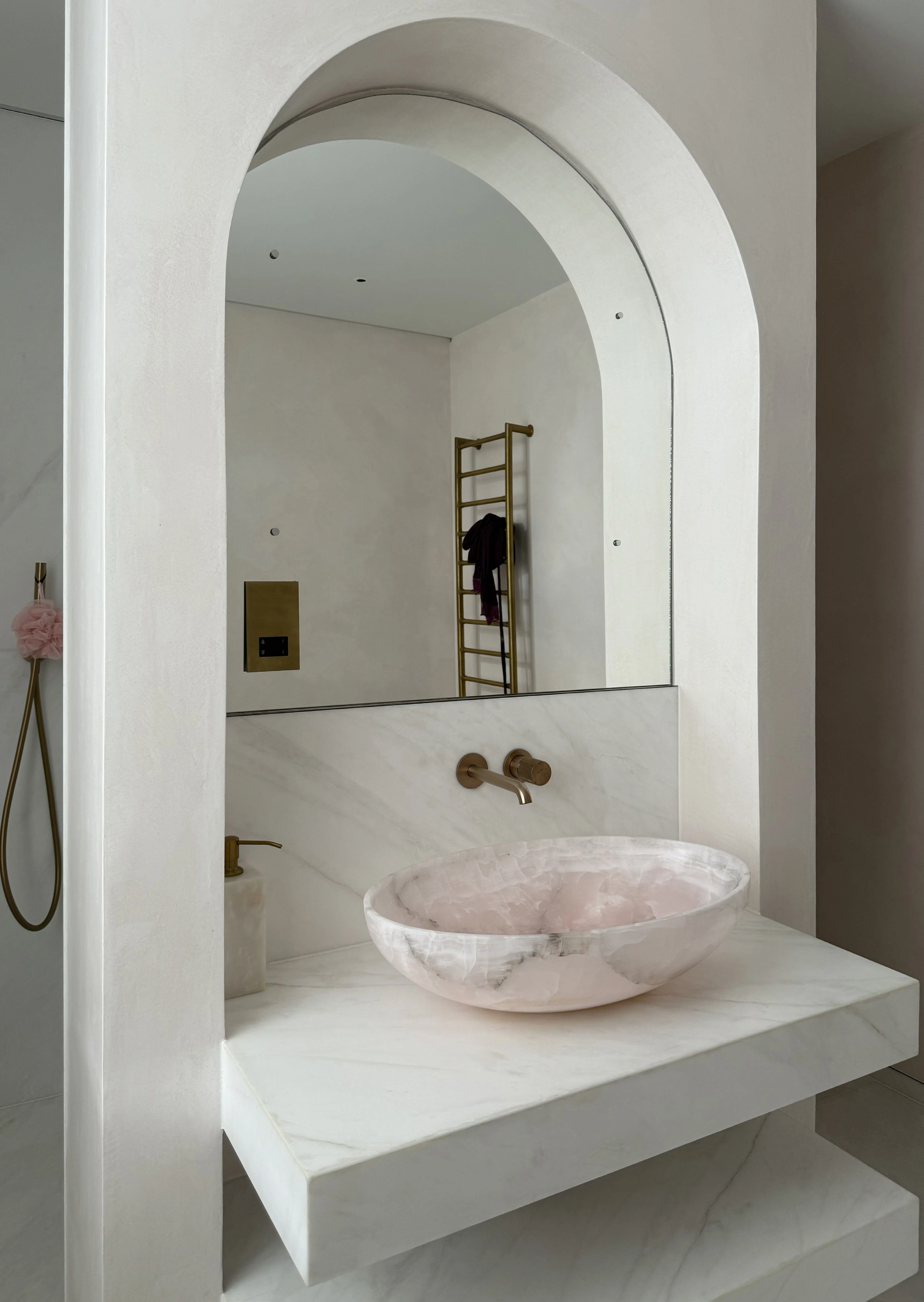 Powder-room vanity with a tall arched mirror, wall-mounted brass taps and a pink stone vessel basin on a white stone shelf.