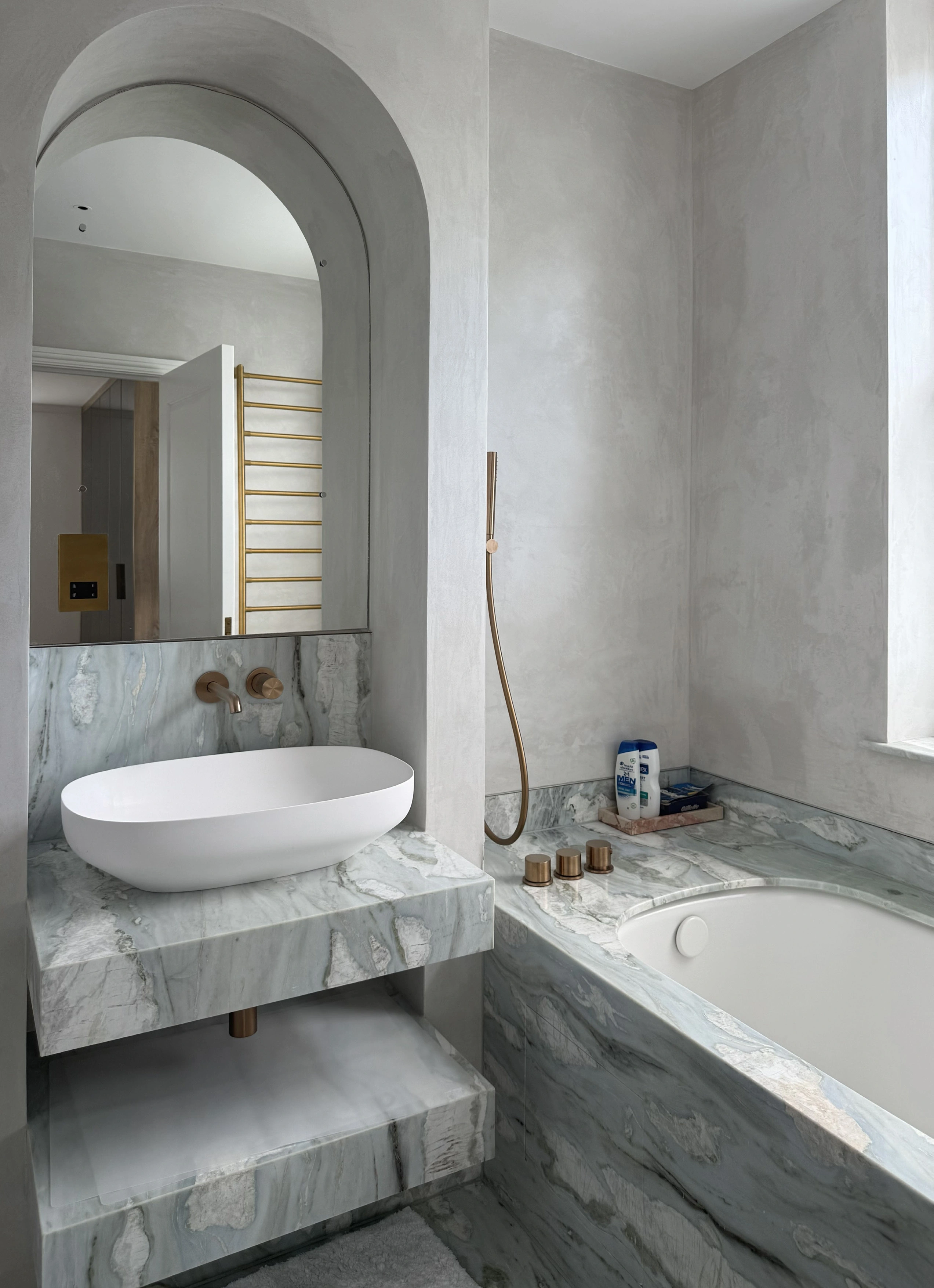 Bathroom with an arched mirror above a white oval basin, grey-veined marble vanity and built-in bath with brass fixtures.