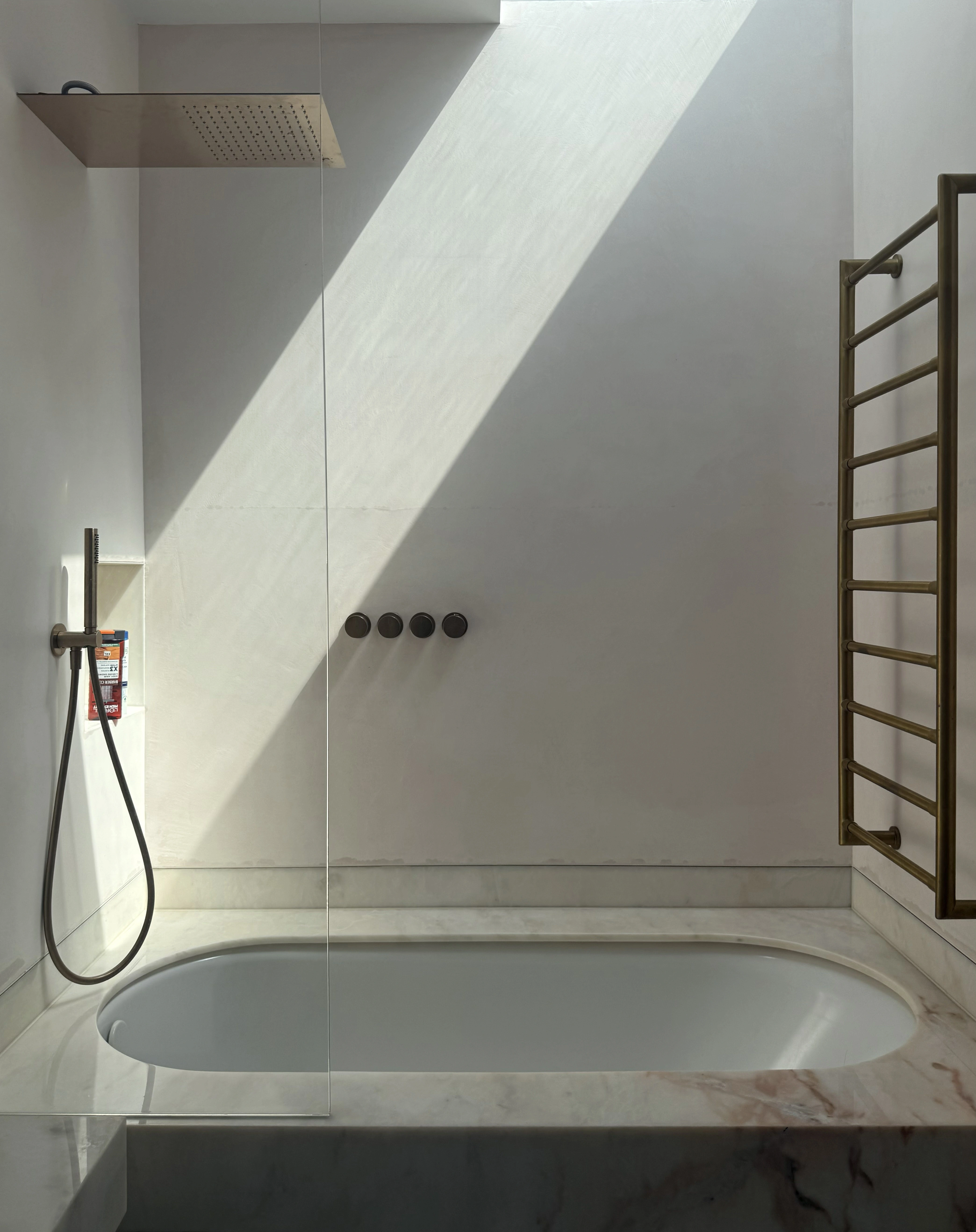 Sunlit bathroom with a built-in bath, frameless glass screen, rain shower and brass heated towel rail.